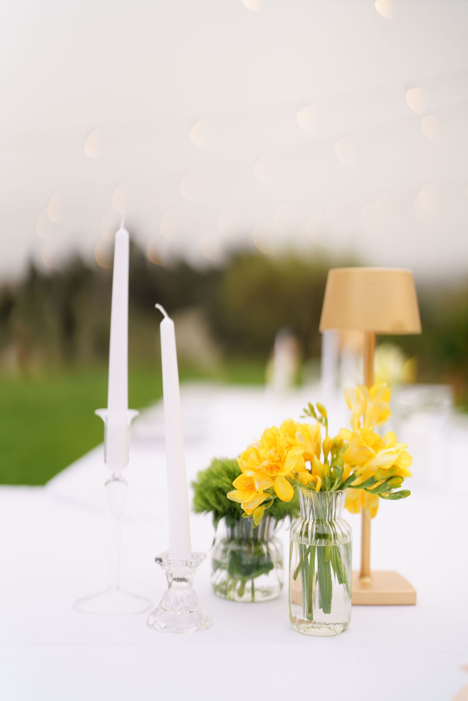 Wedding table decor featuring white candles, yellow flowers in glass vase, and gold table lamp.