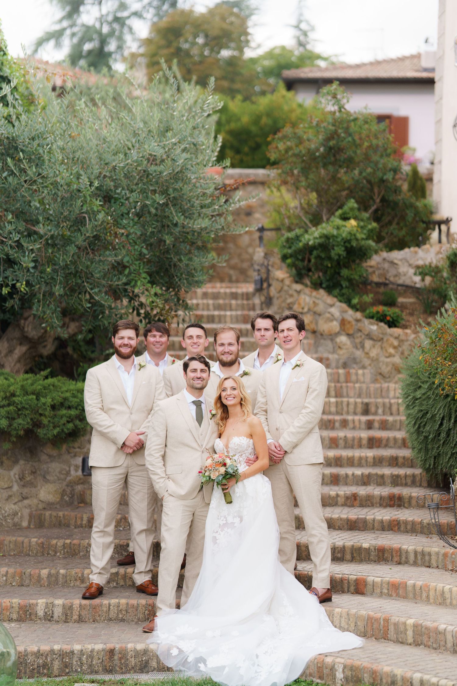 Wedding party in khaki suits and white dress pose on stone steps at outdoor venue.