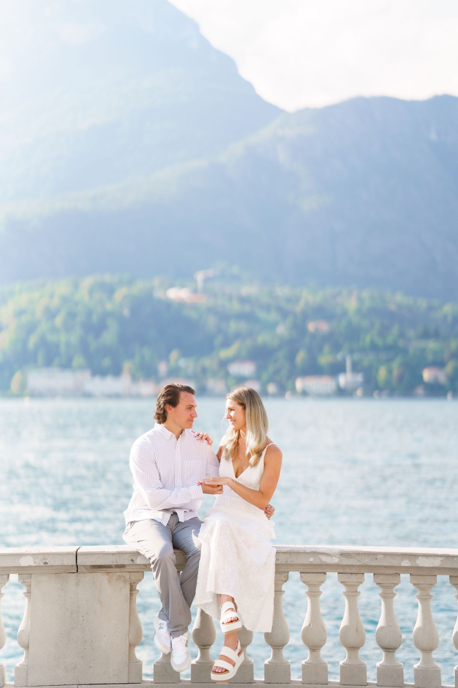 A couple shares a romantic moment on a lakeside balustrade with mountains in the background.