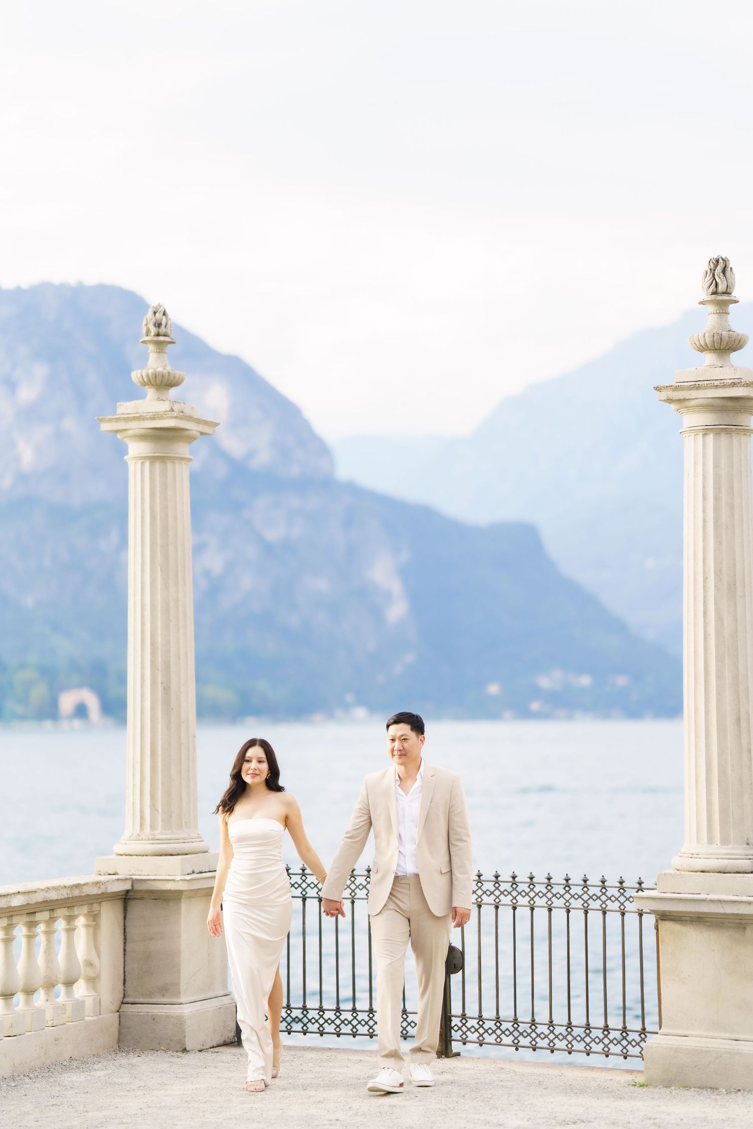 A couple in light colored outfits walks along an elegant seaside terrace with white columns and mountain views.