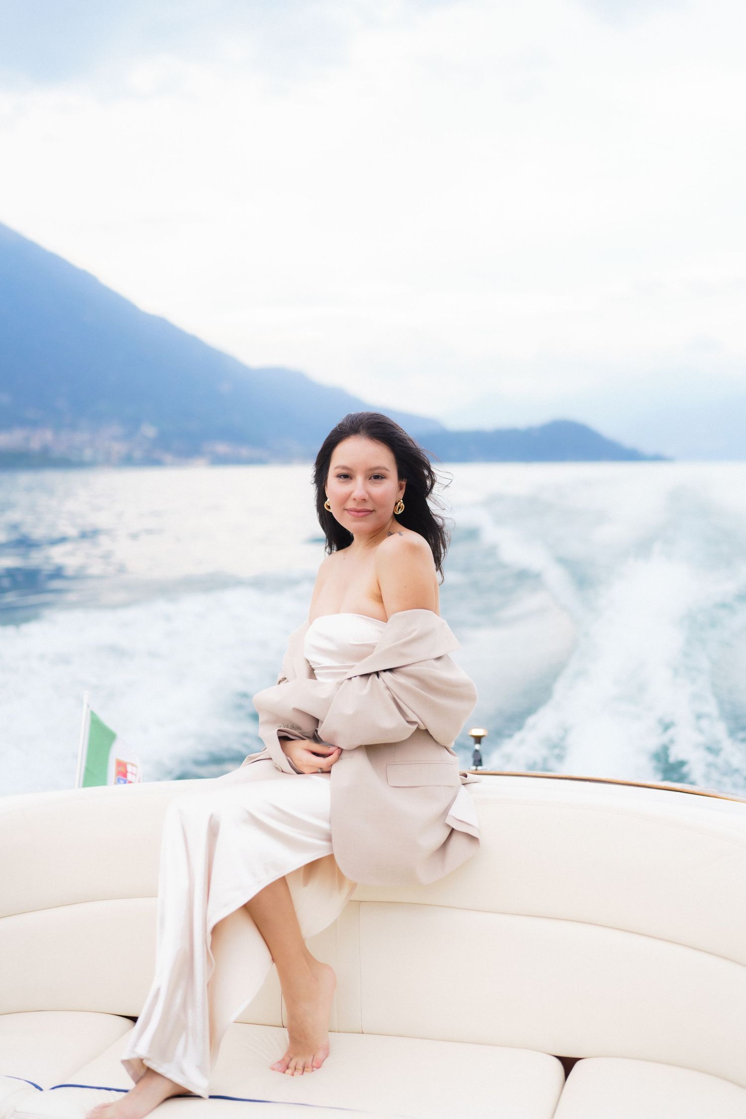 A person in beige dress relaxes on a luxury yacht with coastal mountains and blue water in the background.