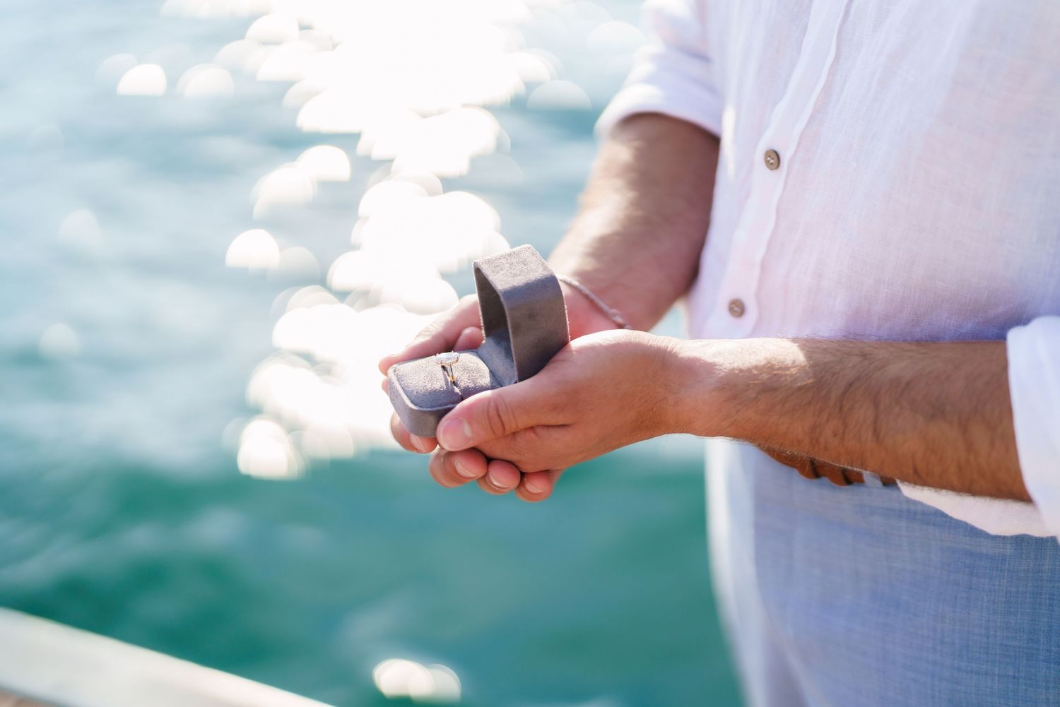 Man in white shirt holding open ring box over sparkling ocean water, preparing for a marriage proposal.