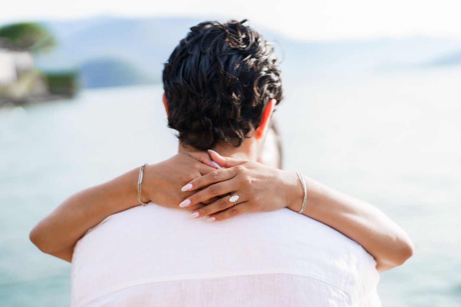 Couple embracing outdoors near water, woman with short dark hair wearing white, diamond ring visible on hand.