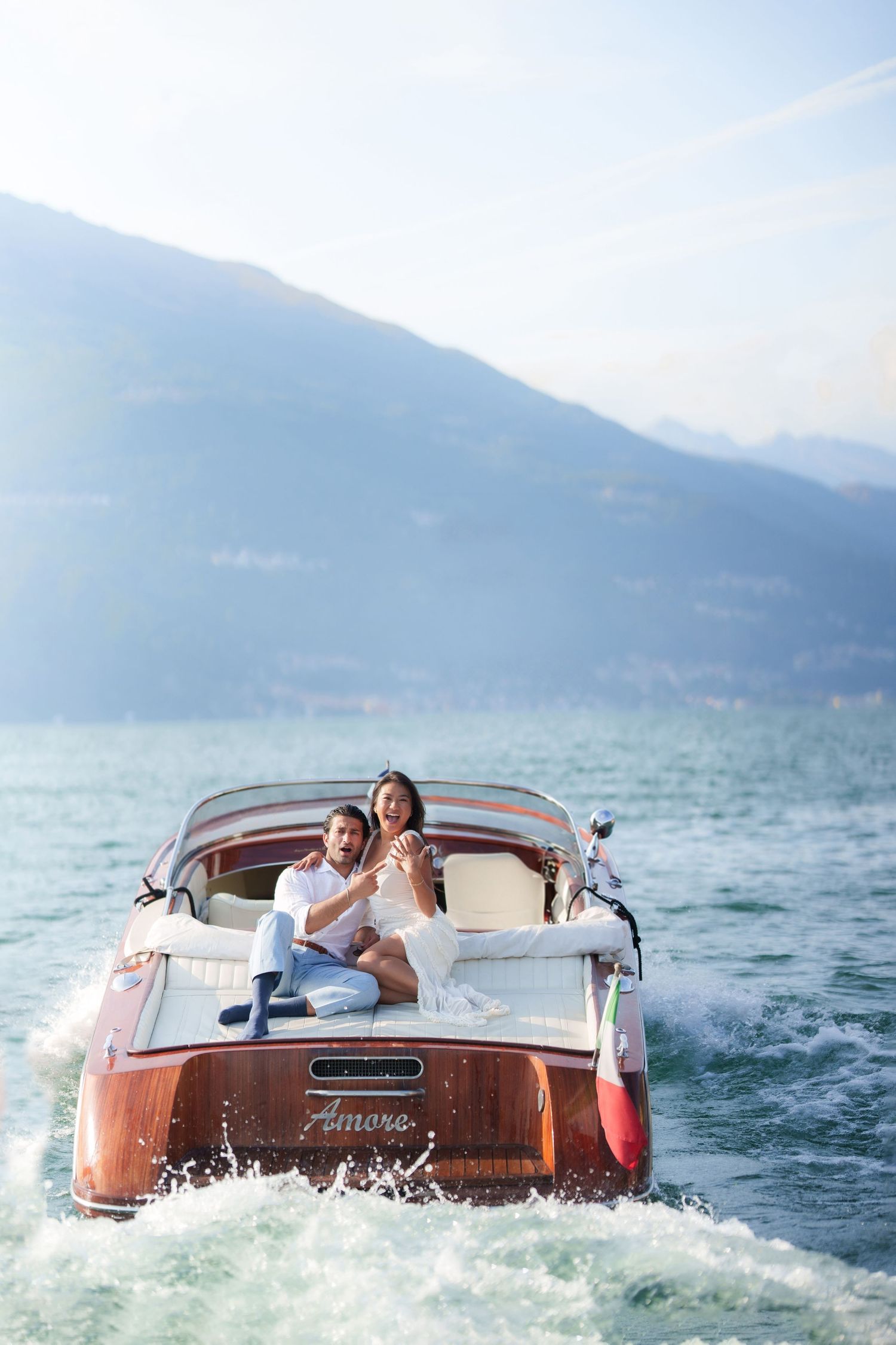 Couple riding a classic wooden Riva speedboat named Amore on a scenic alpine lake with mountains in the background.