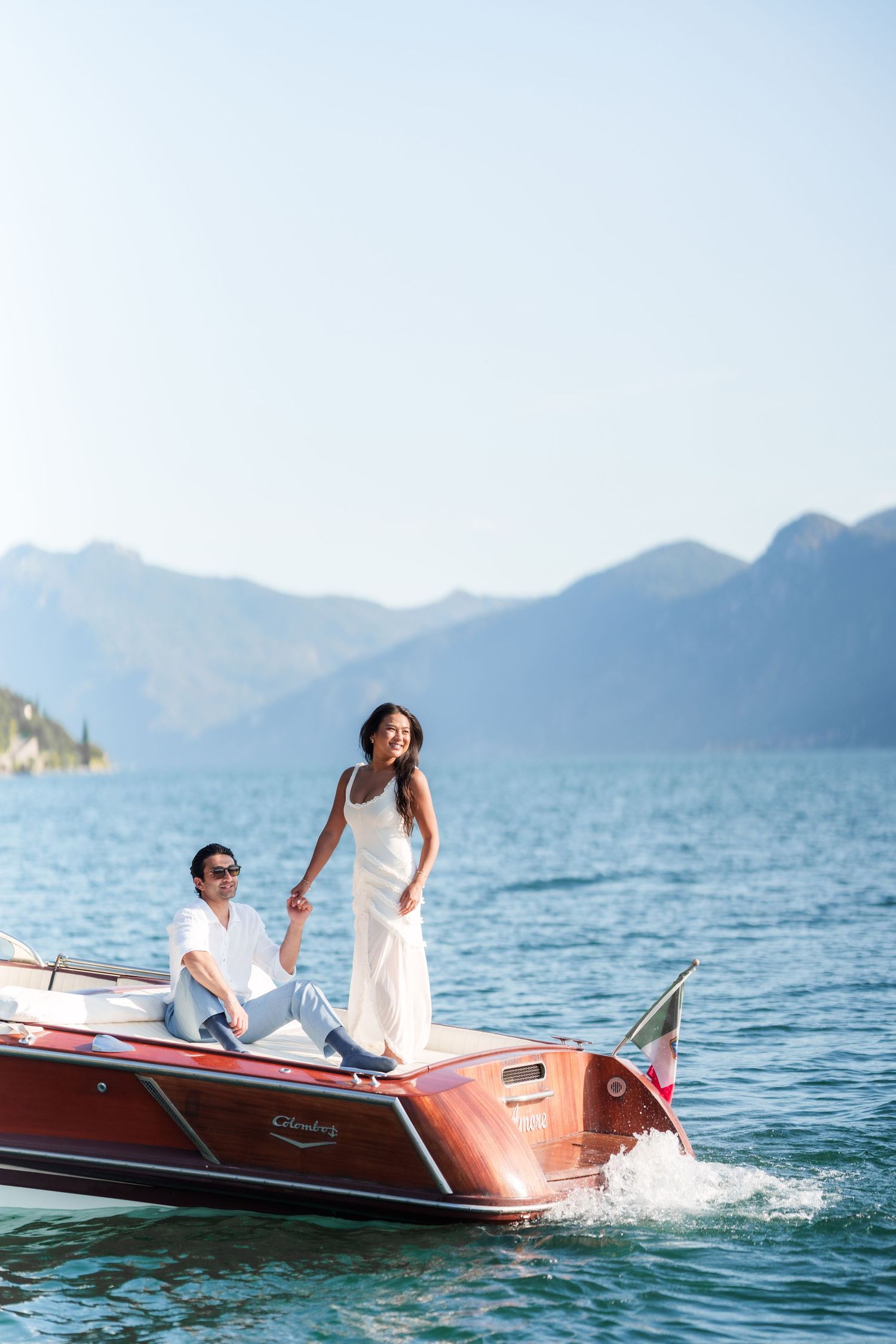 Couple in white outfits riding a classic wooden speedboat on a serene alpine lake with mountain backdrop.