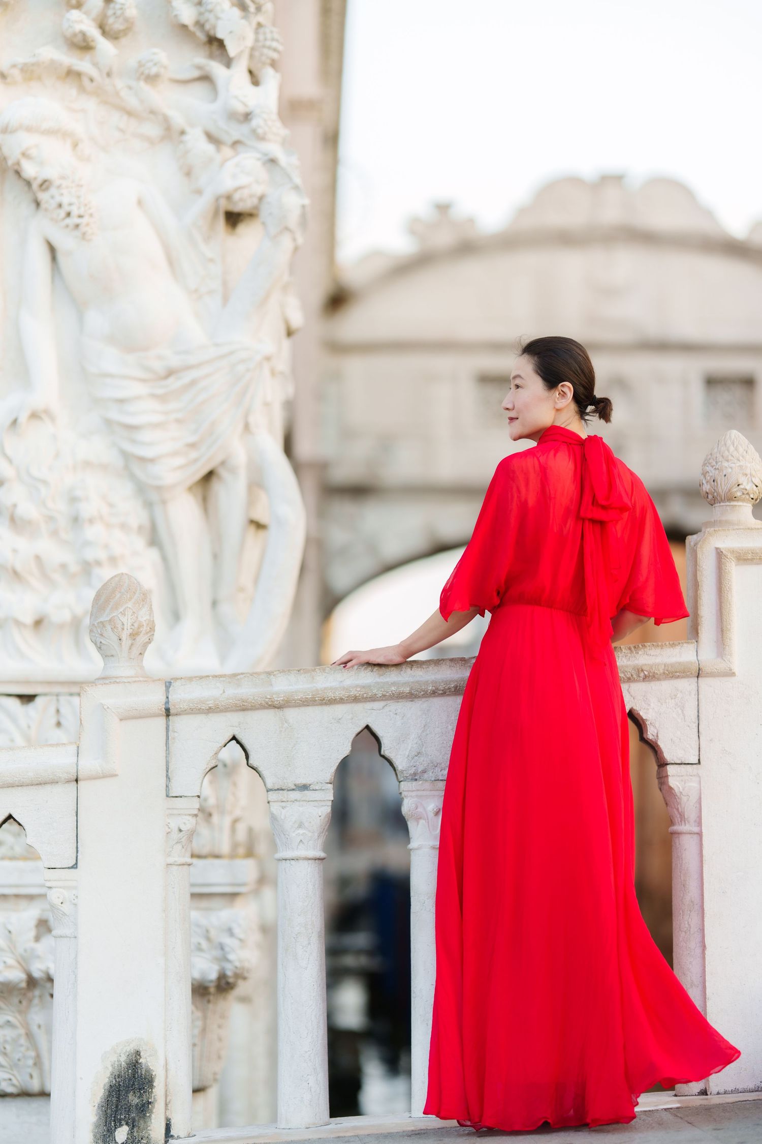 Woman in elegant red gown standing on marble balcony beside classical sculpture in European setting.