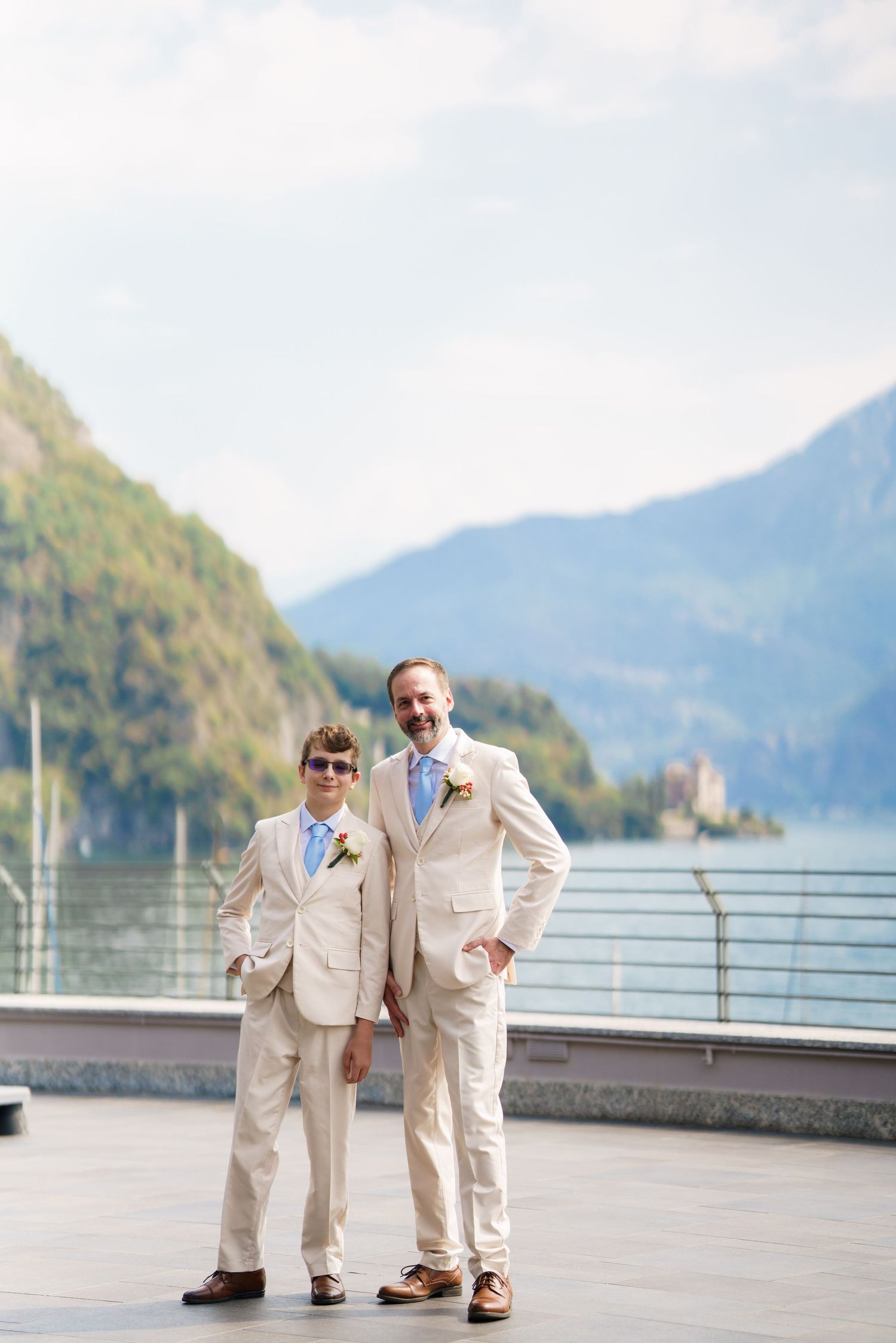 Father and son in matching cream suits pose on a lakeside terrace with mountain backdrop at a wedding.