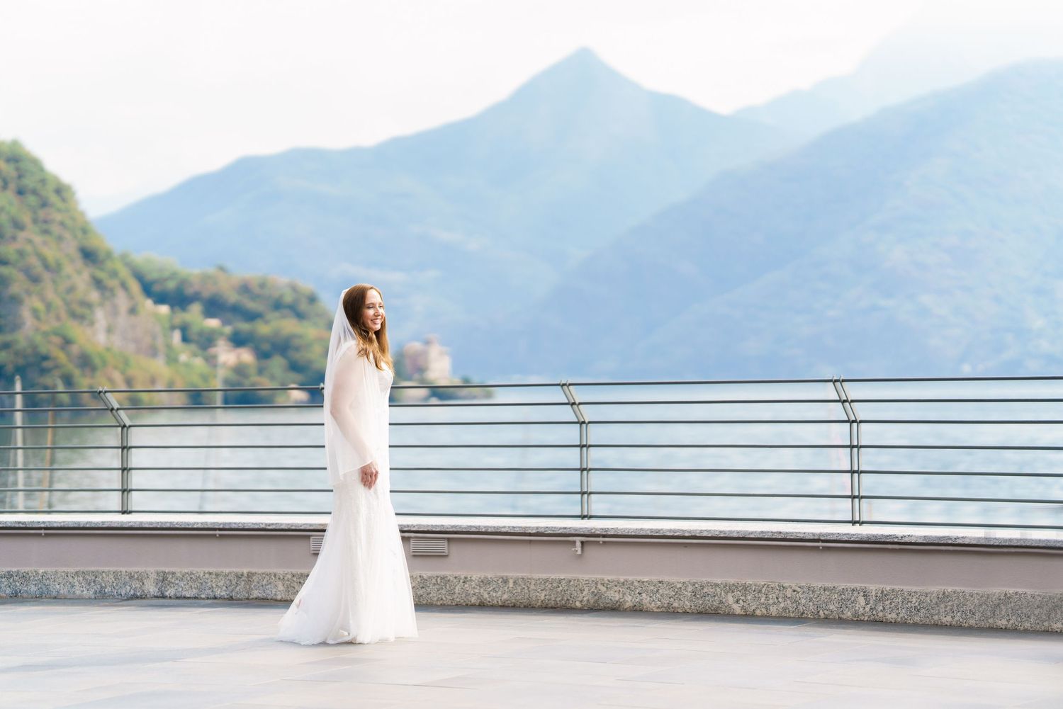 Bride in white gown poses on lakeside terrace with mountain backdrop at Lake Como wedding.