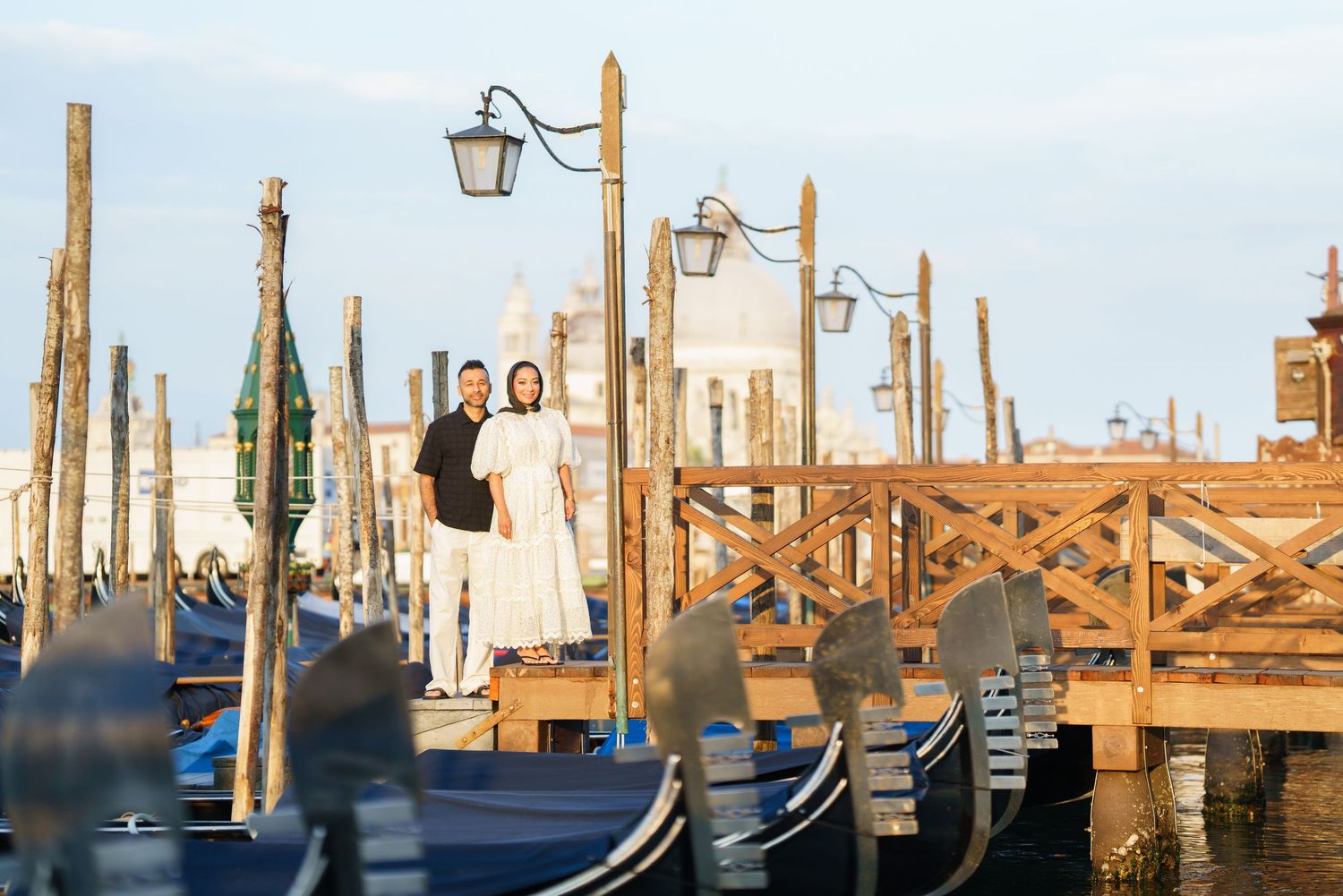 Couple in white attire posing on a Venice gondola dock with iconic wooden poles and blue gondolas.