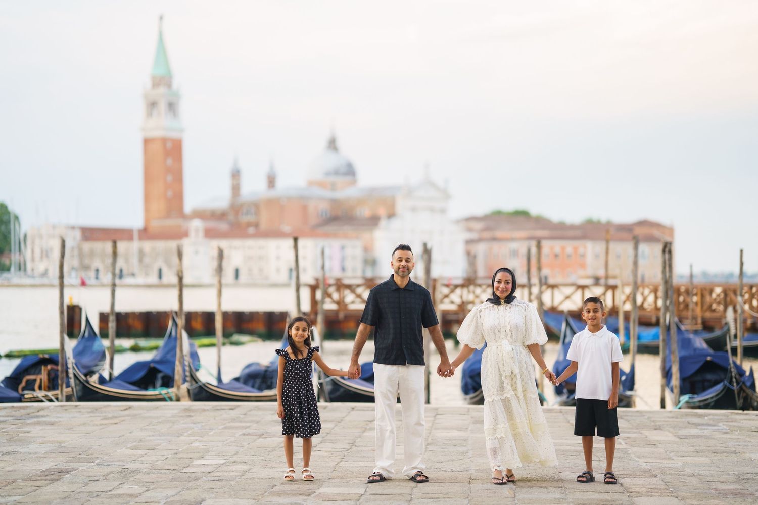 Family of four holding hands on Venice waterfront with gondolas and San Giorgio Maggiore church in background.