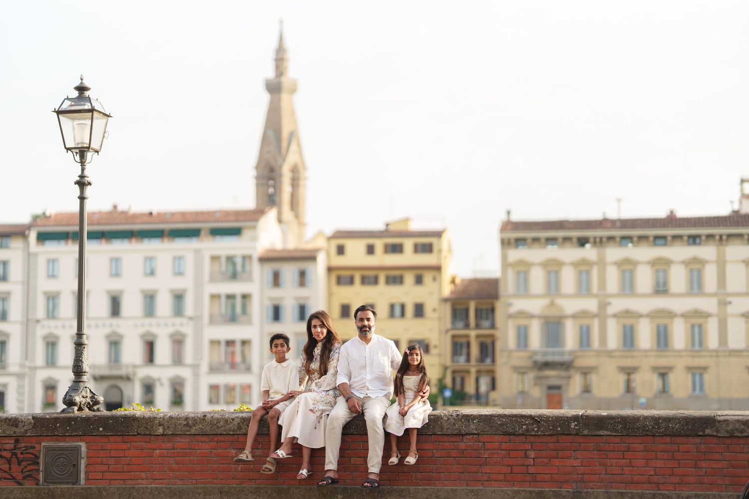 Family of four sitting on a brick wall in Florence, Italy, with colorful buildings and a church tower in the background.