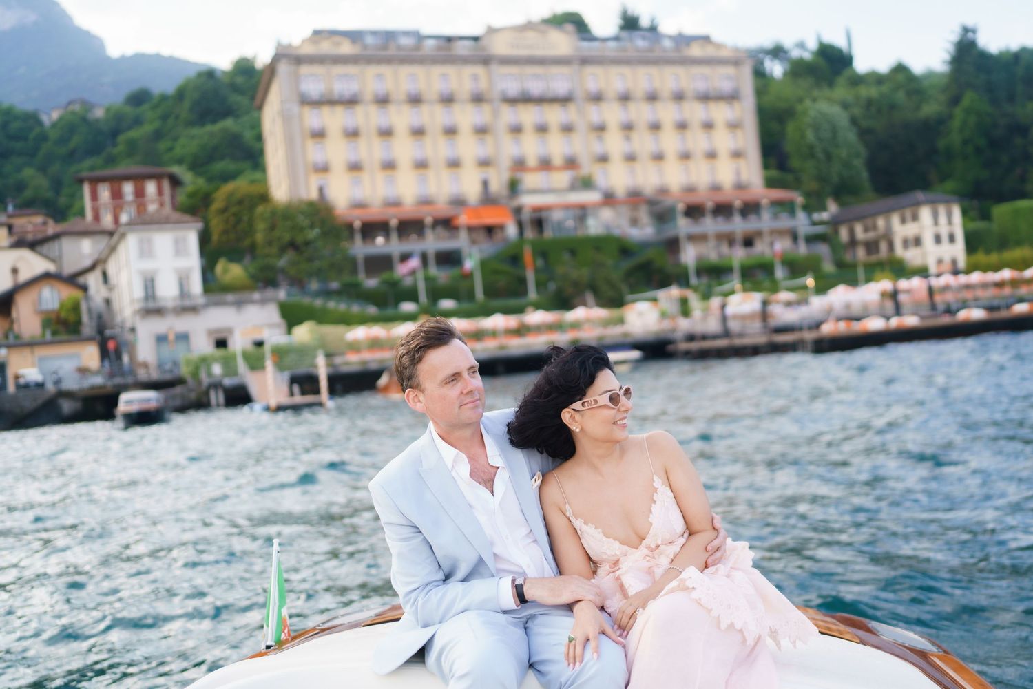 Couple sitting together on a wooden boat with Lake Como and a grand hotel in the background.