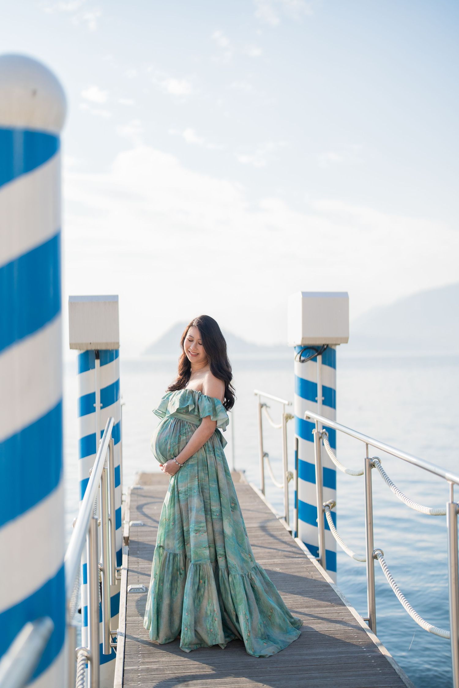 Pregnant woman in teal off-shoulder gown stands on a pier with blue striped posts, serene lake and mountains behind her.