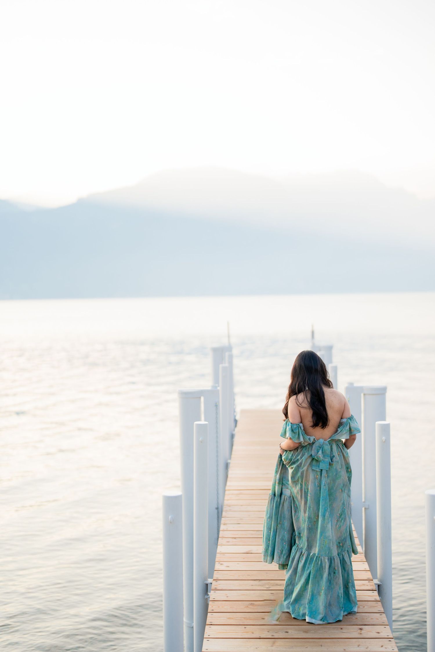 Woman in teal floral maxi dress standing on wooden pier overlooking calm lake with misty mountains.