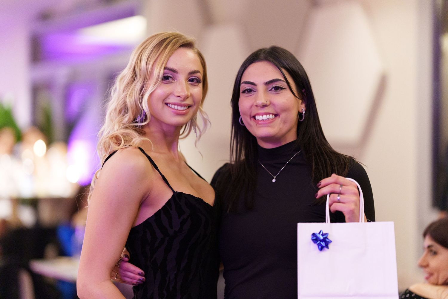 Two women in black outfits smiling together at an event, one holding a white gift bag with blue ribbon.