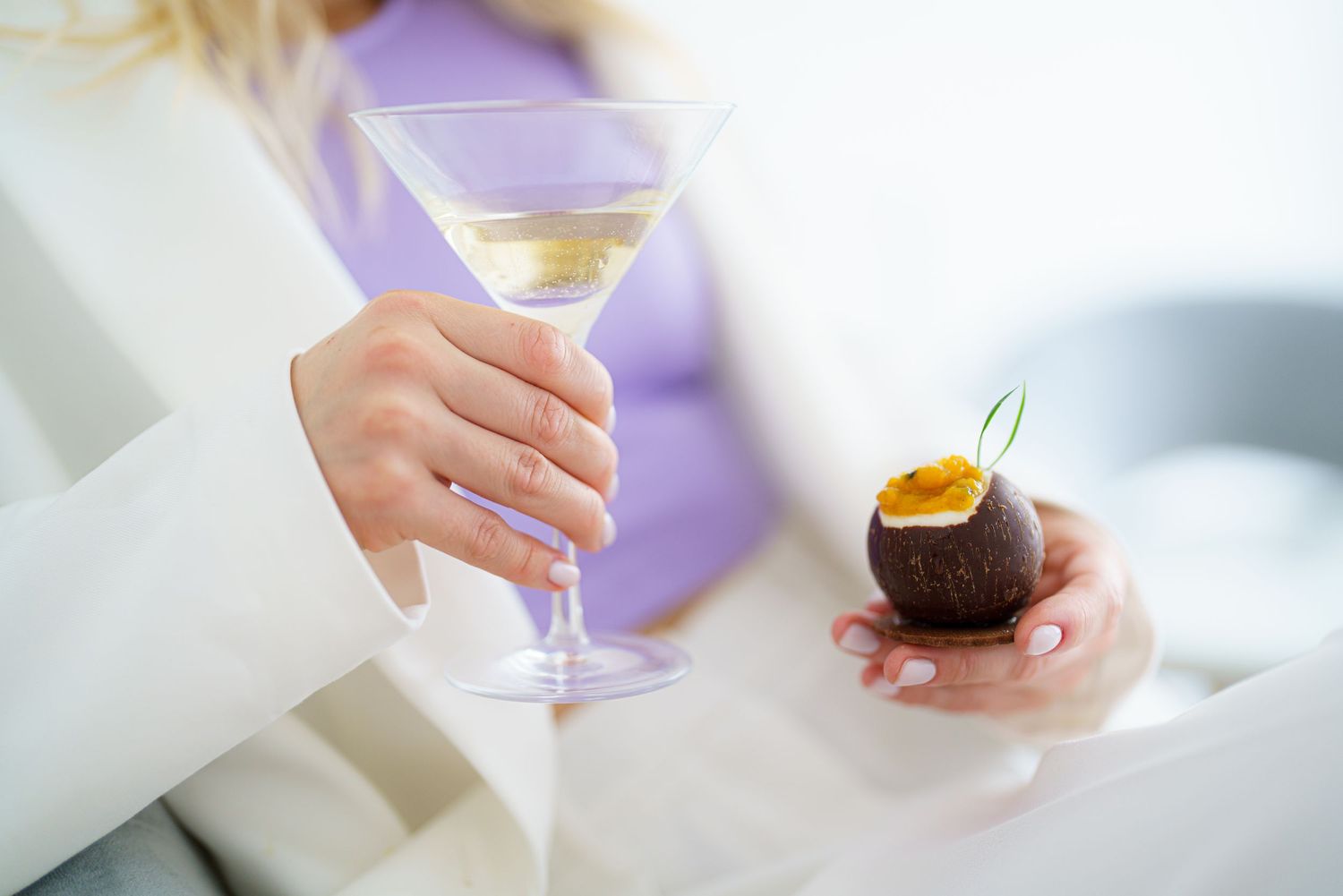 Woman holding a martini glass and a chocolate dessert with passion fruit at an elegant event.