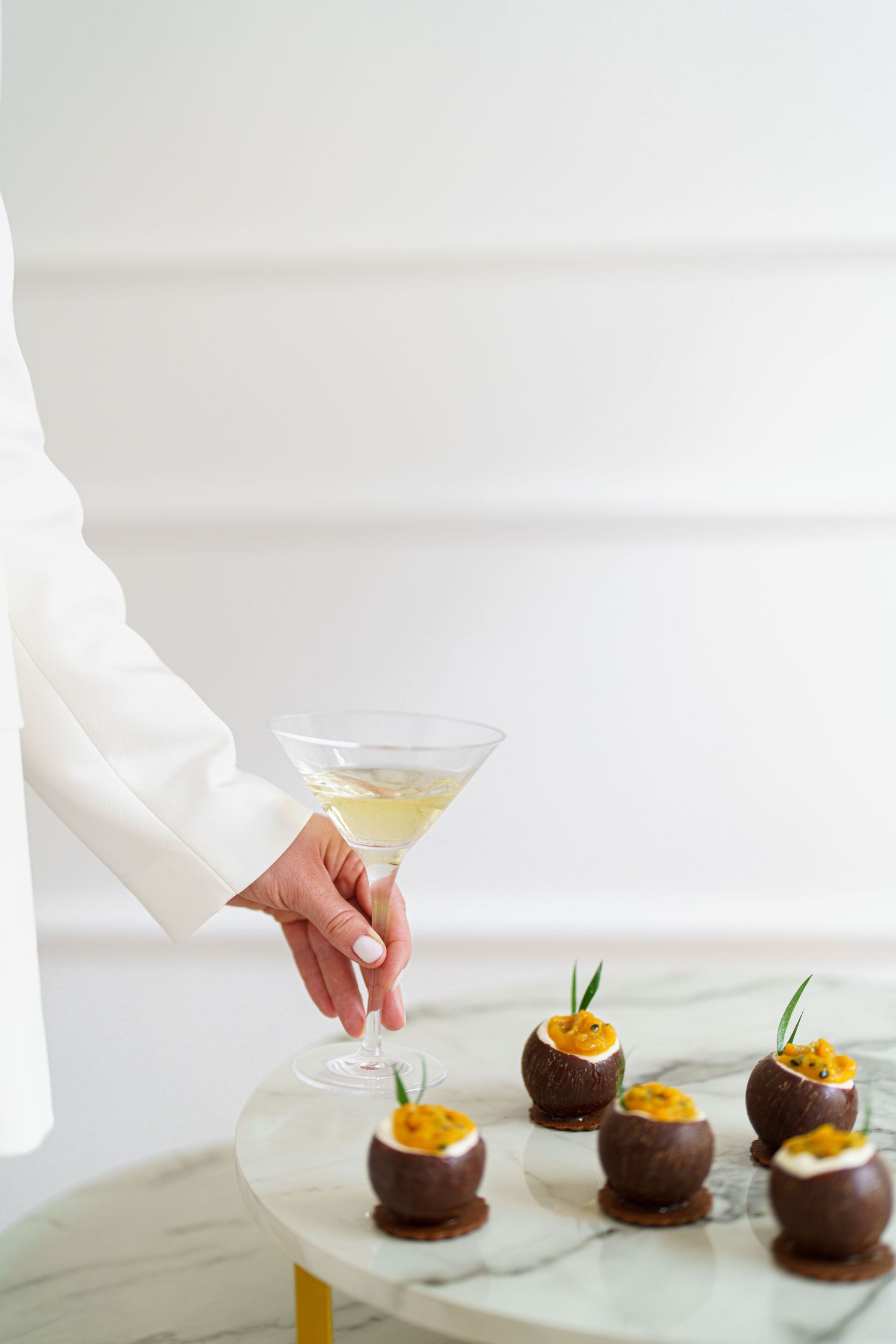 A hand holding a martini glass near elegant chocolate sphere appetizers topped with orange garnish on a marble table.