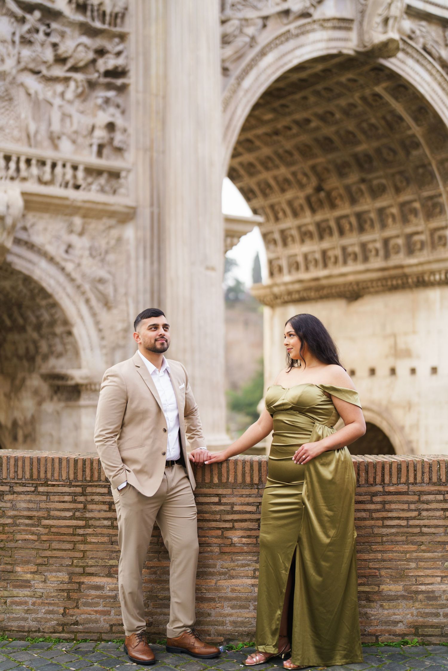 Couple in elegant attire poses before ancient Roman arch, man in tan suit, woman in olive green gown.