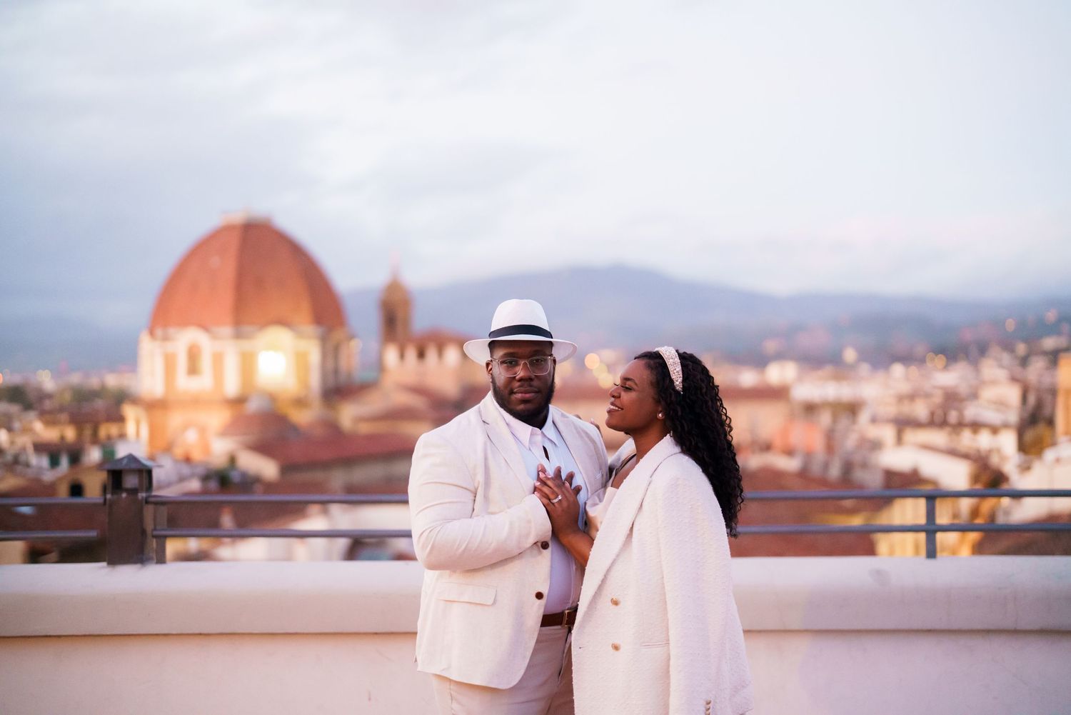Couple in white attire posing on a rooftop terrace with Florence Cathedral dome and cityscape at dusk.
