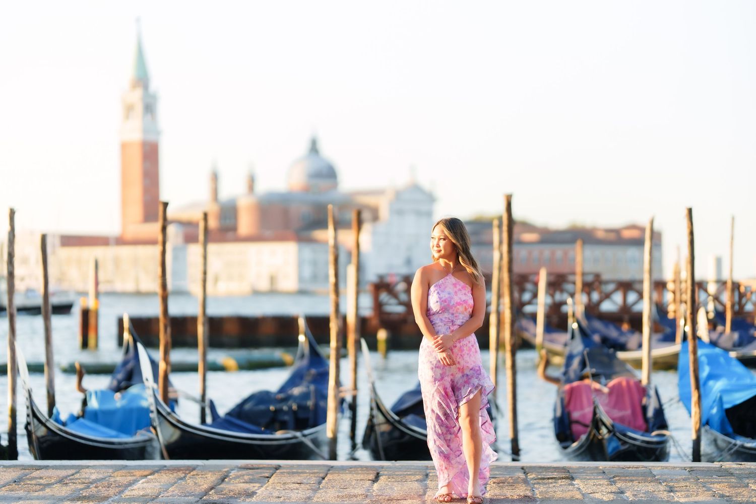 Woman in pink dress posing by Venice gondolas with San Giorgio Maggiore church in background at sunset.