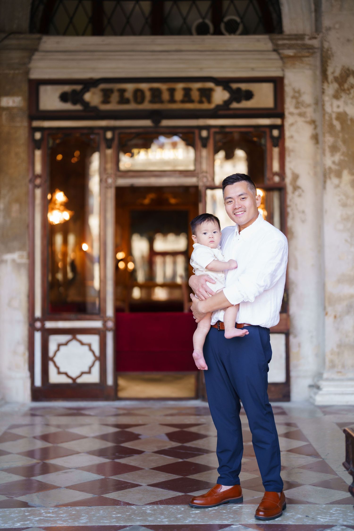 A smiling man holds a baby outside Cafe Florian in Venice, Italy, standing on marble floors.