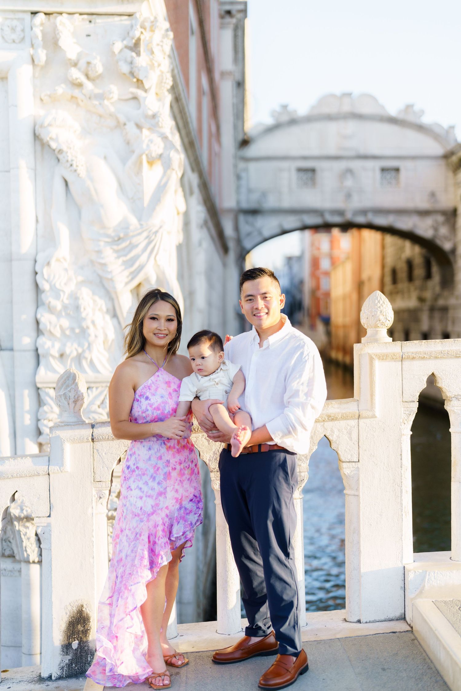A smiling family of three poses on a Venice bridge near the Bridge of Sighs, with a canal in the background.