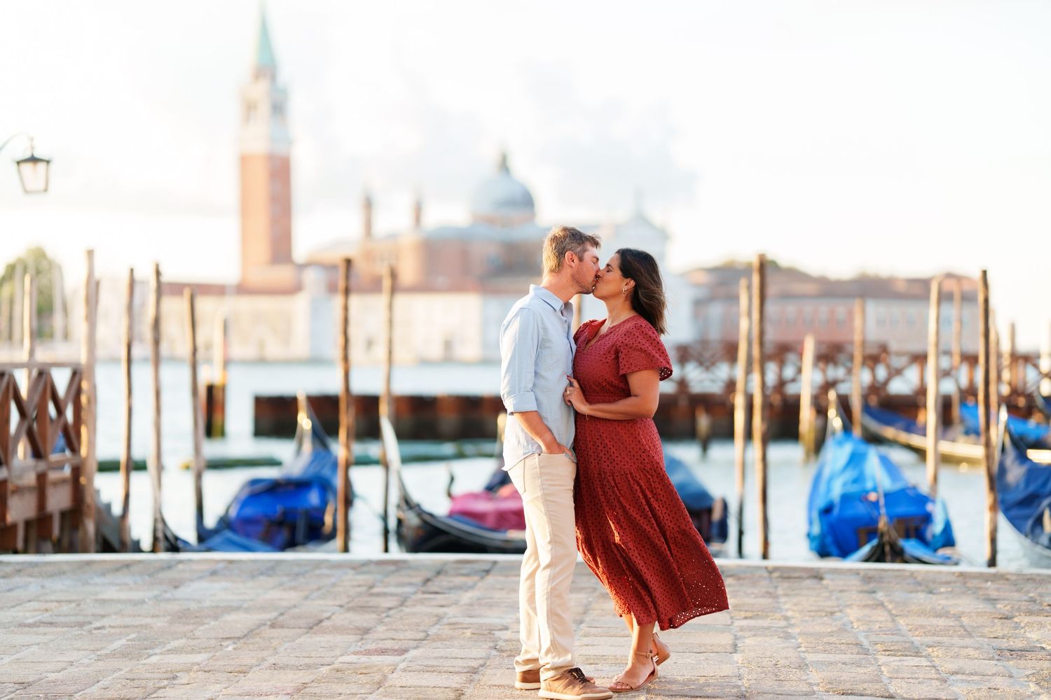 Couple sharing a romantic kiss by Venice gondola docks with San Giorgio Maggiore church in the background.