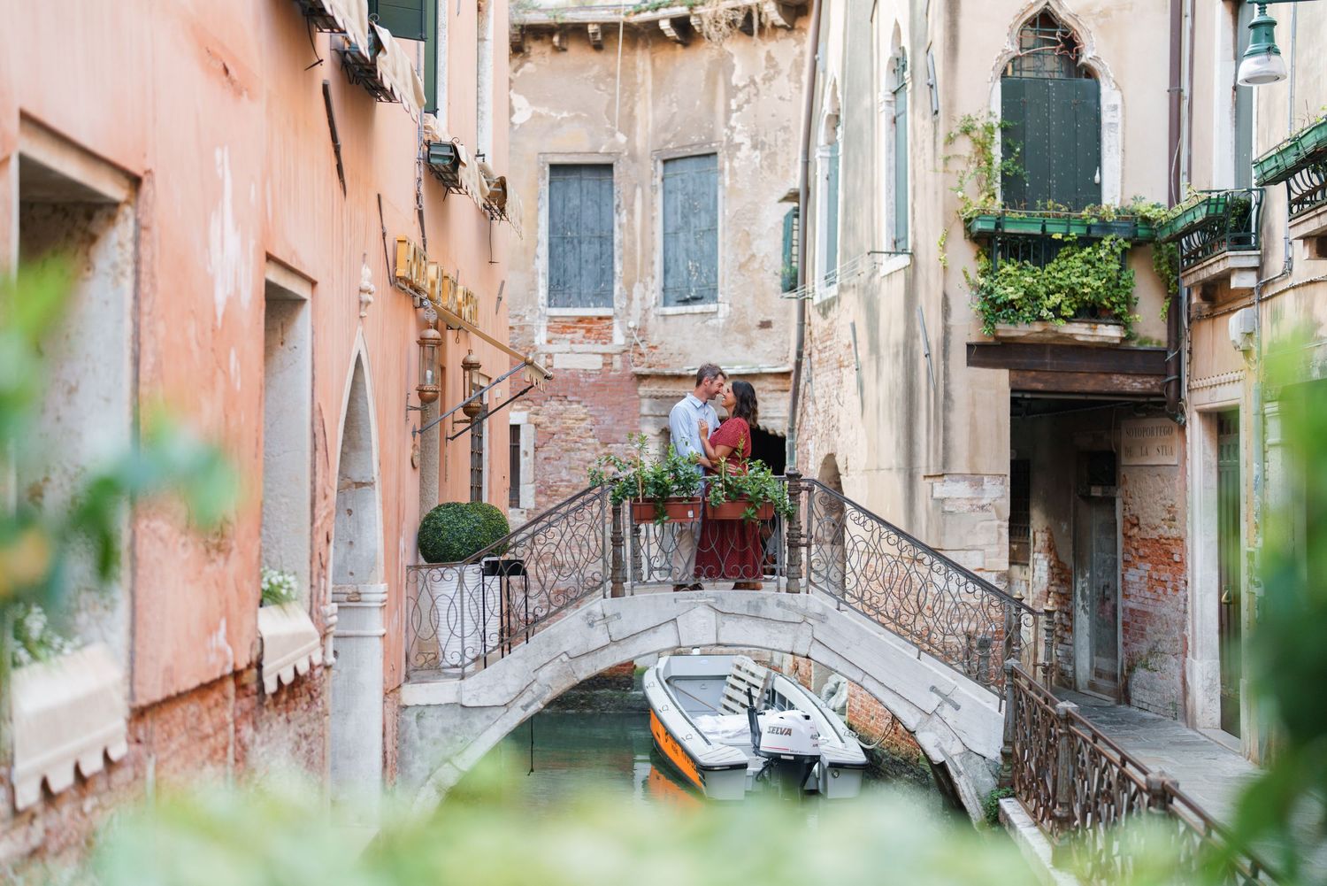 Couple embracing on a stone bridge over a Venice canal, surrounded by colorful historic buildings and lush greenery.