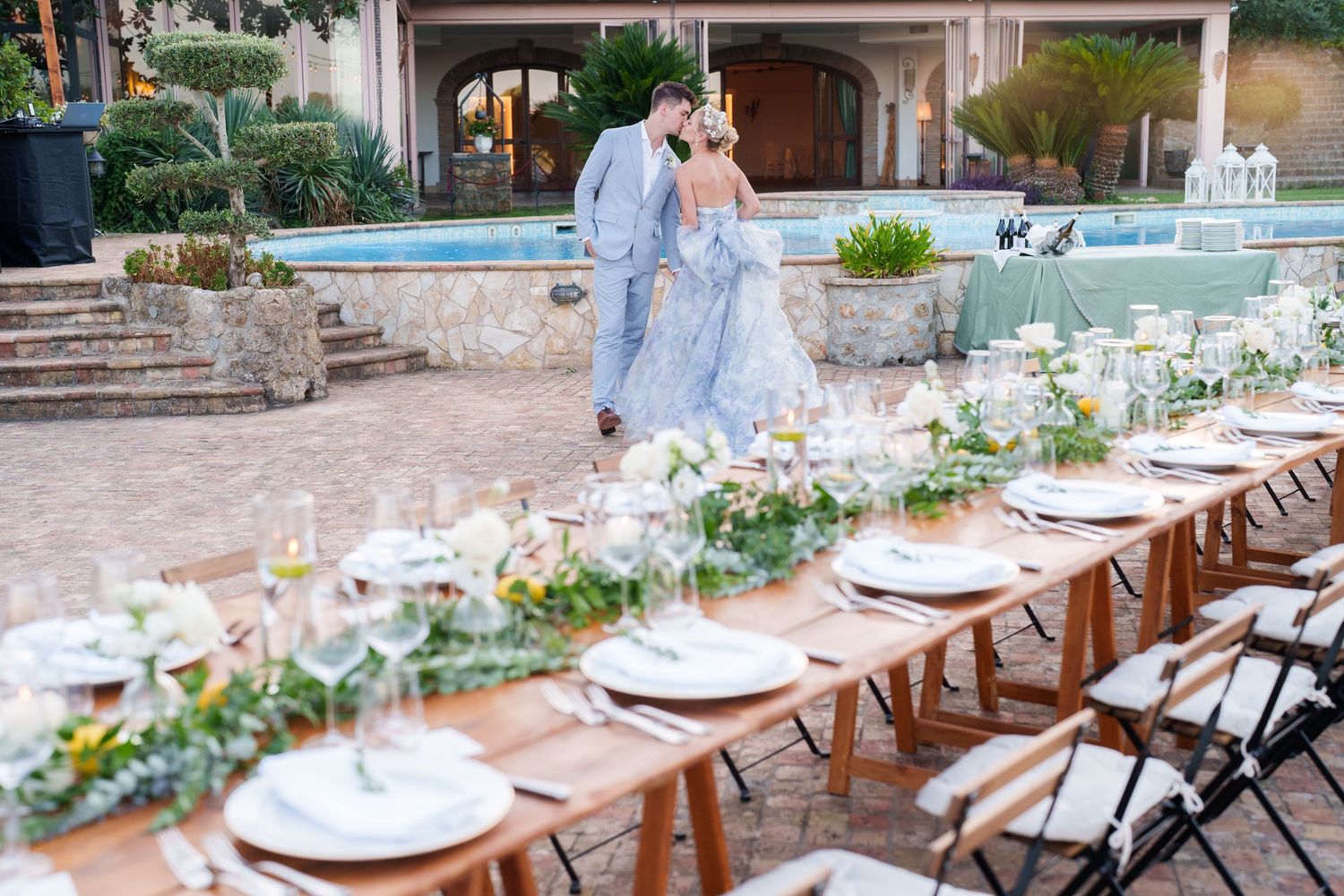 Couple in blue wedding attire walks past elegantly set outdoor reception tables with floral greenery garlands at luxury venue.
