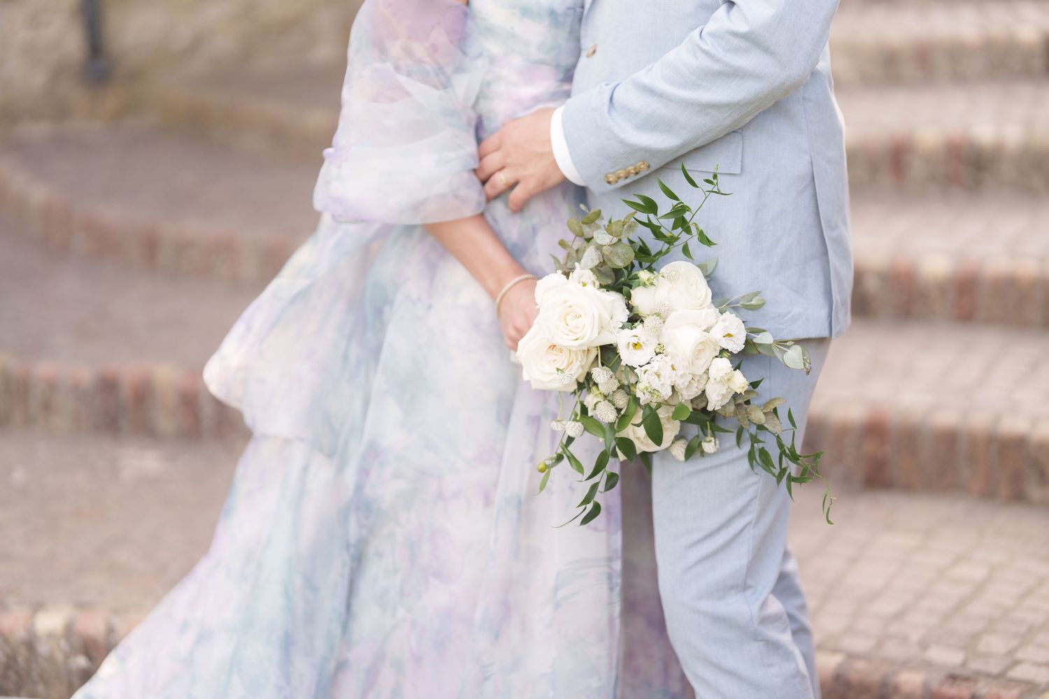 Bride in pastel watercolor gown and groom in light blue suit holding white rose bouquet at wedding.