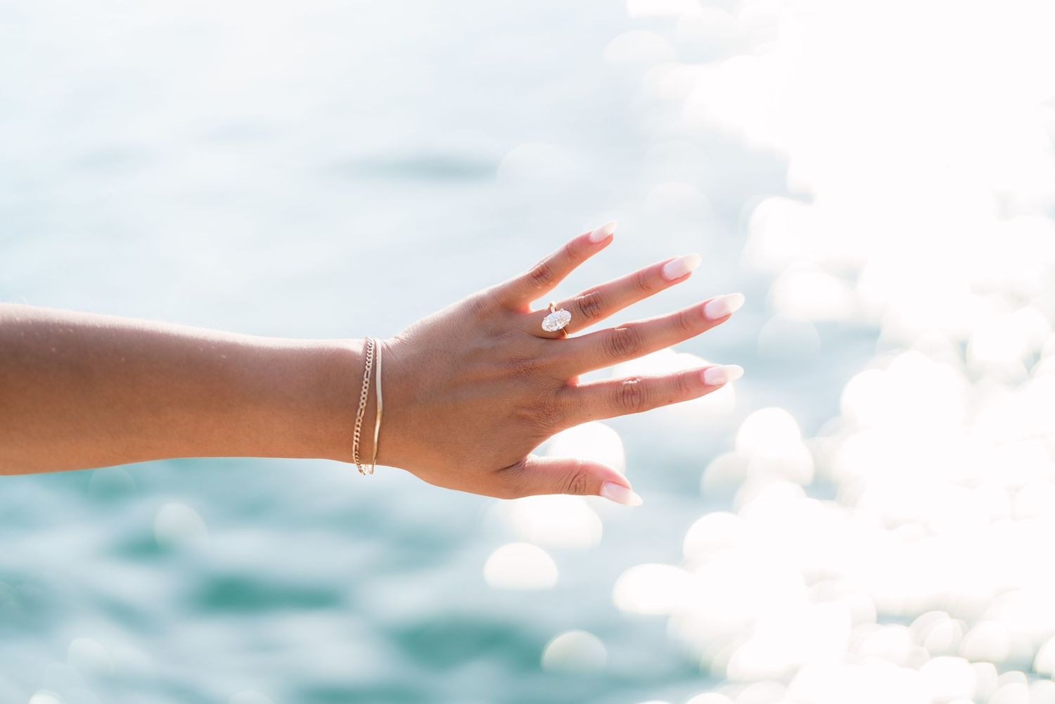 Woman's hand adorned with a diamond ring and gold bracelet raised against sparkling blue ocean water.