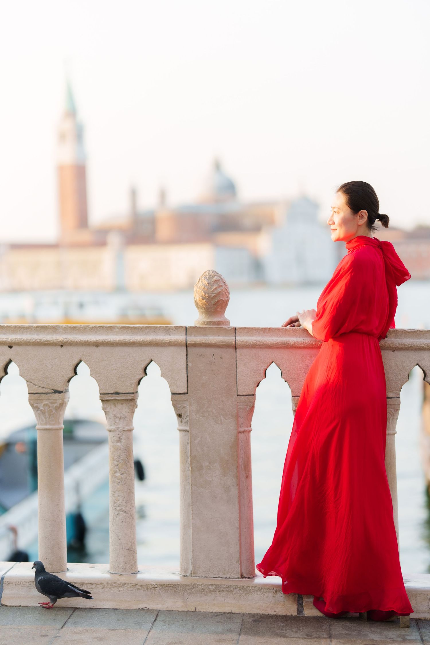 Woman in red dress standing on Venice balcony overlooking the lagoon with San Giorgio Maggiore in background.