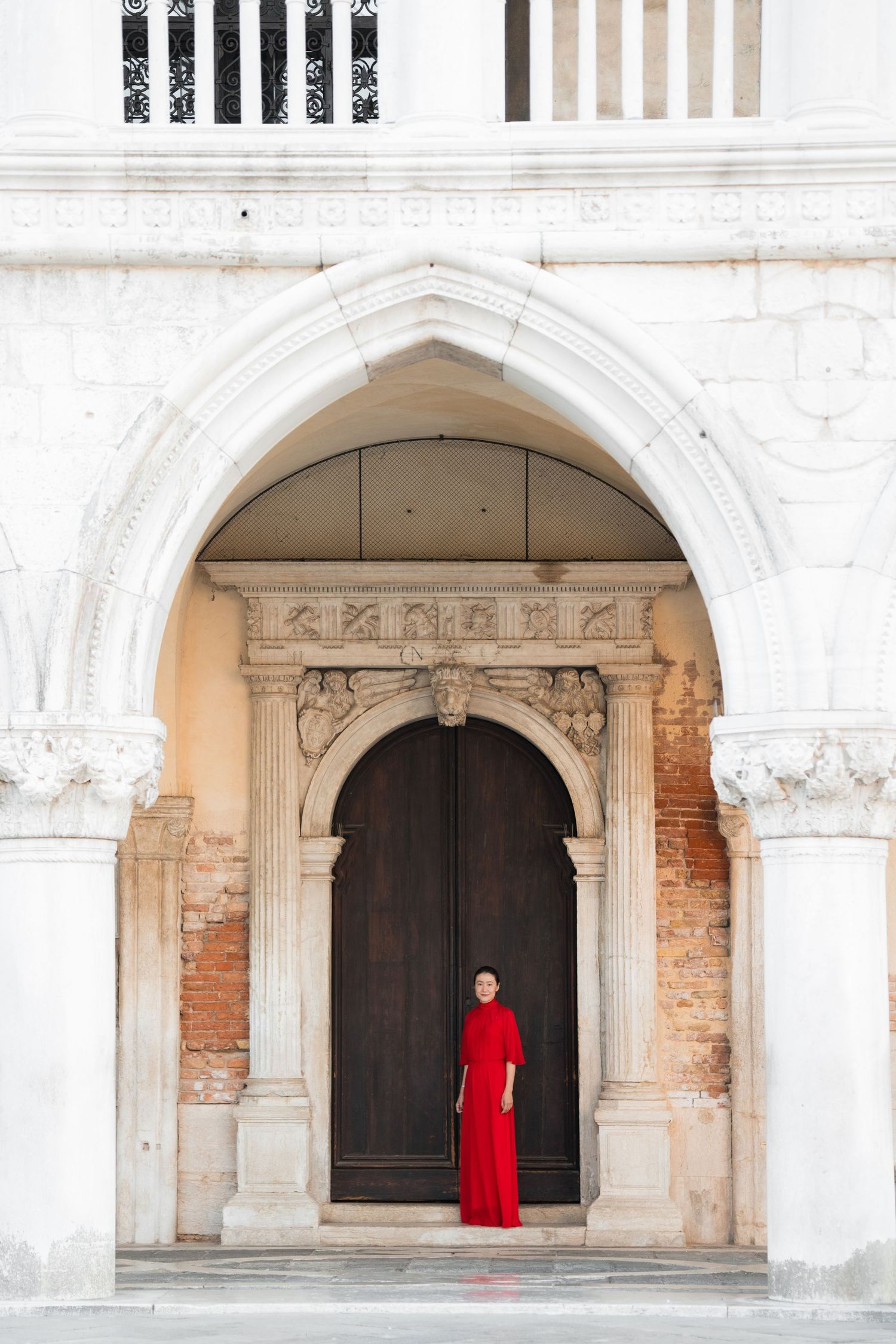 Woman in red dress standing in ornate Gothic arch doorway of historic Venetian building with marble columns.