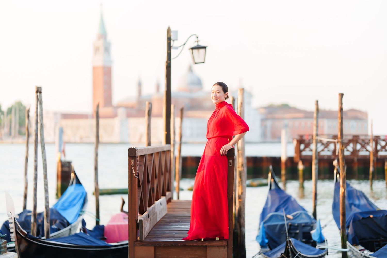 Woman in red dress posing by Venice gondolas with San Giorgio Maggiore church in background.