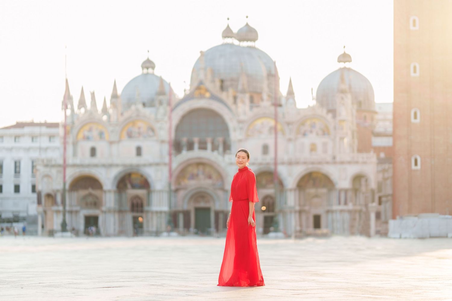 Woman in red gown stands in St. Mark's Square, Venice, with the iconic Basilica di San Marco in the background.