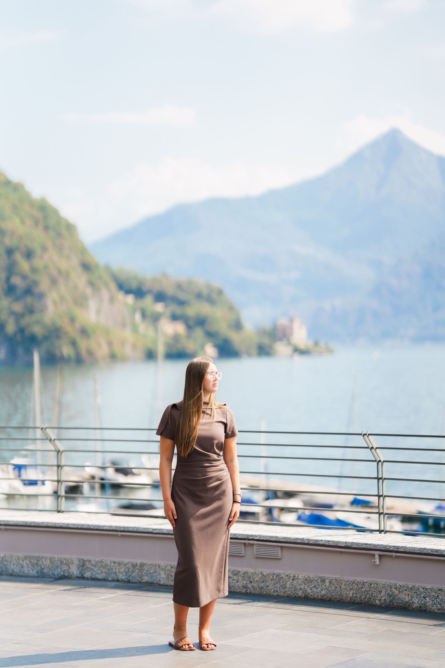 Woman in brown dress standing on lakeside terrace with mountain and marina views on a sunny day.