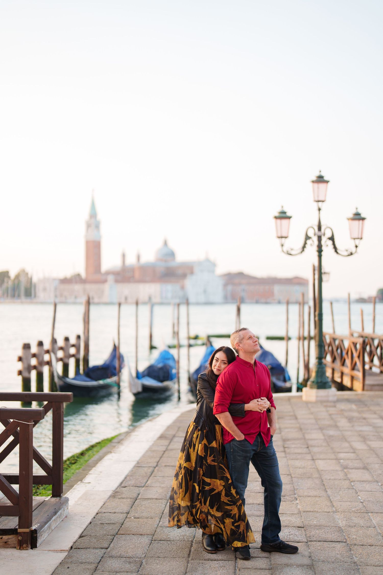 A couple embraces on a misty morning along Venice's waterfront with gondolas and ornate lampposts in the background.