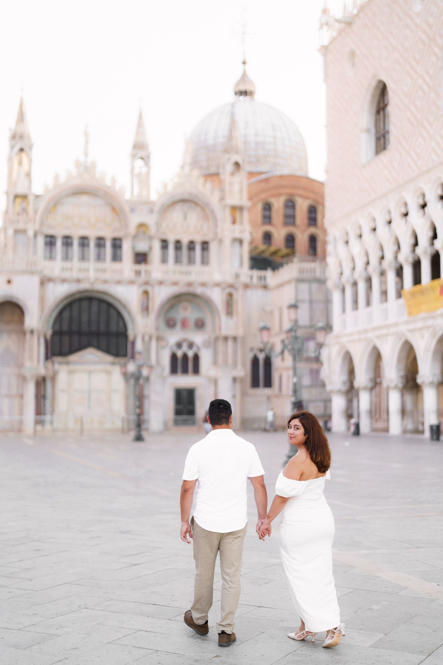 Couple walking through historic St. Mark's Square in Venice with ornate basilica and architecture in background.