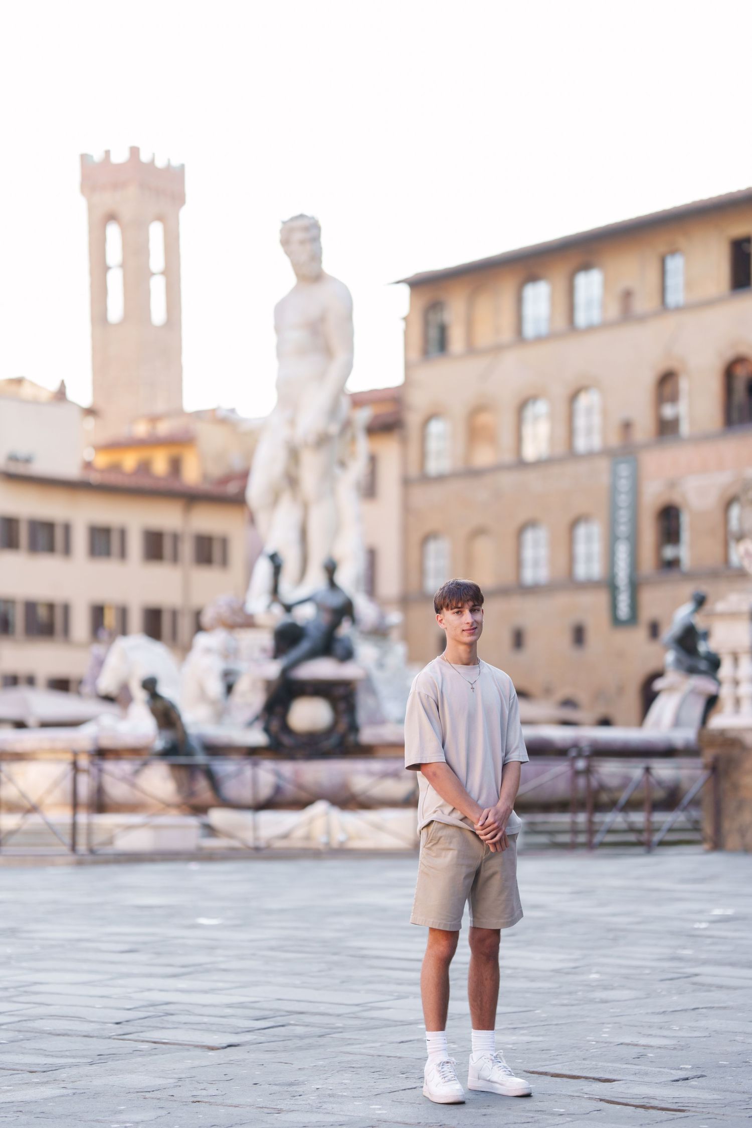 A person in casual attire stands in a historic Italian plaza with stone architecture and towers in the background.