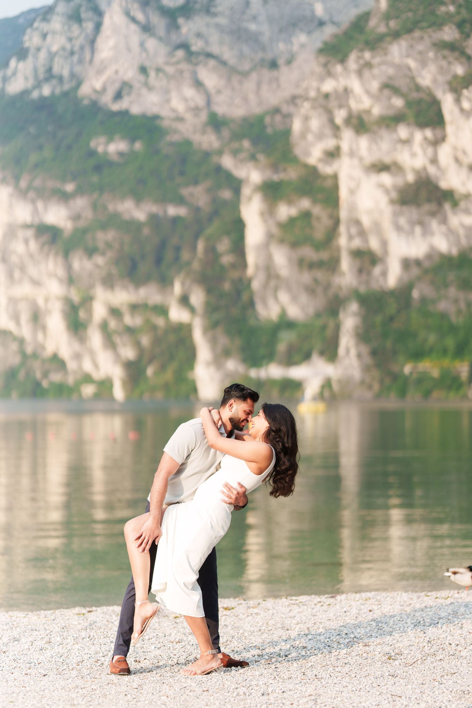 A romantic couple embraces on a rocky beach shore with dramatic mountain cliffs and a serene lake in the background.
