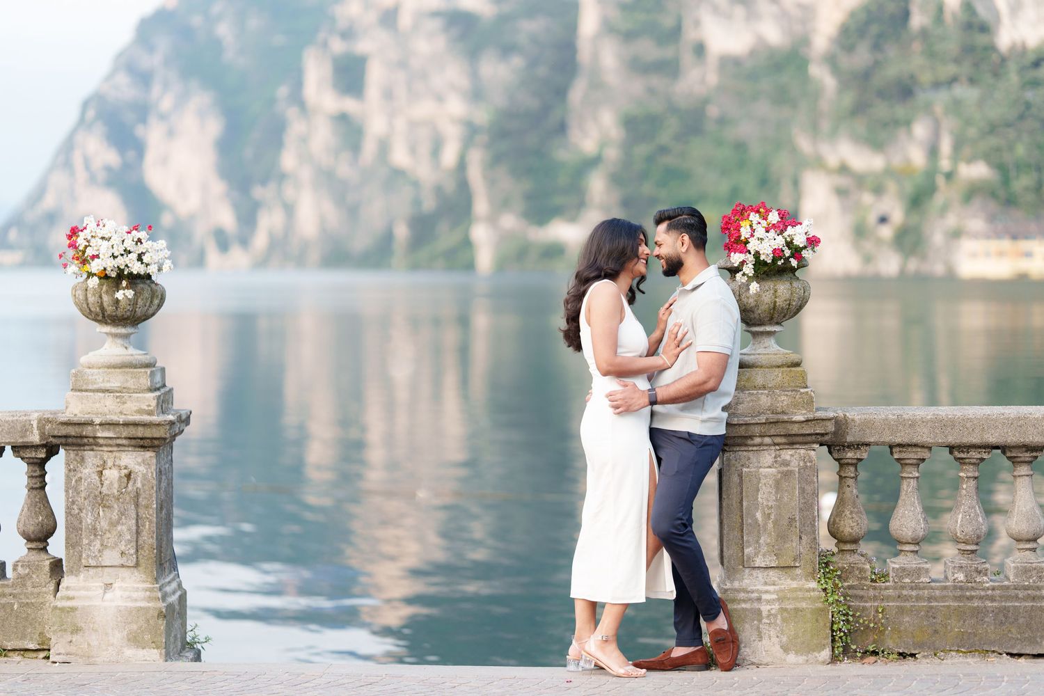 Romantic couple embraces on ornate stone balcony overlooking serene lake with mountain cliffs in background.