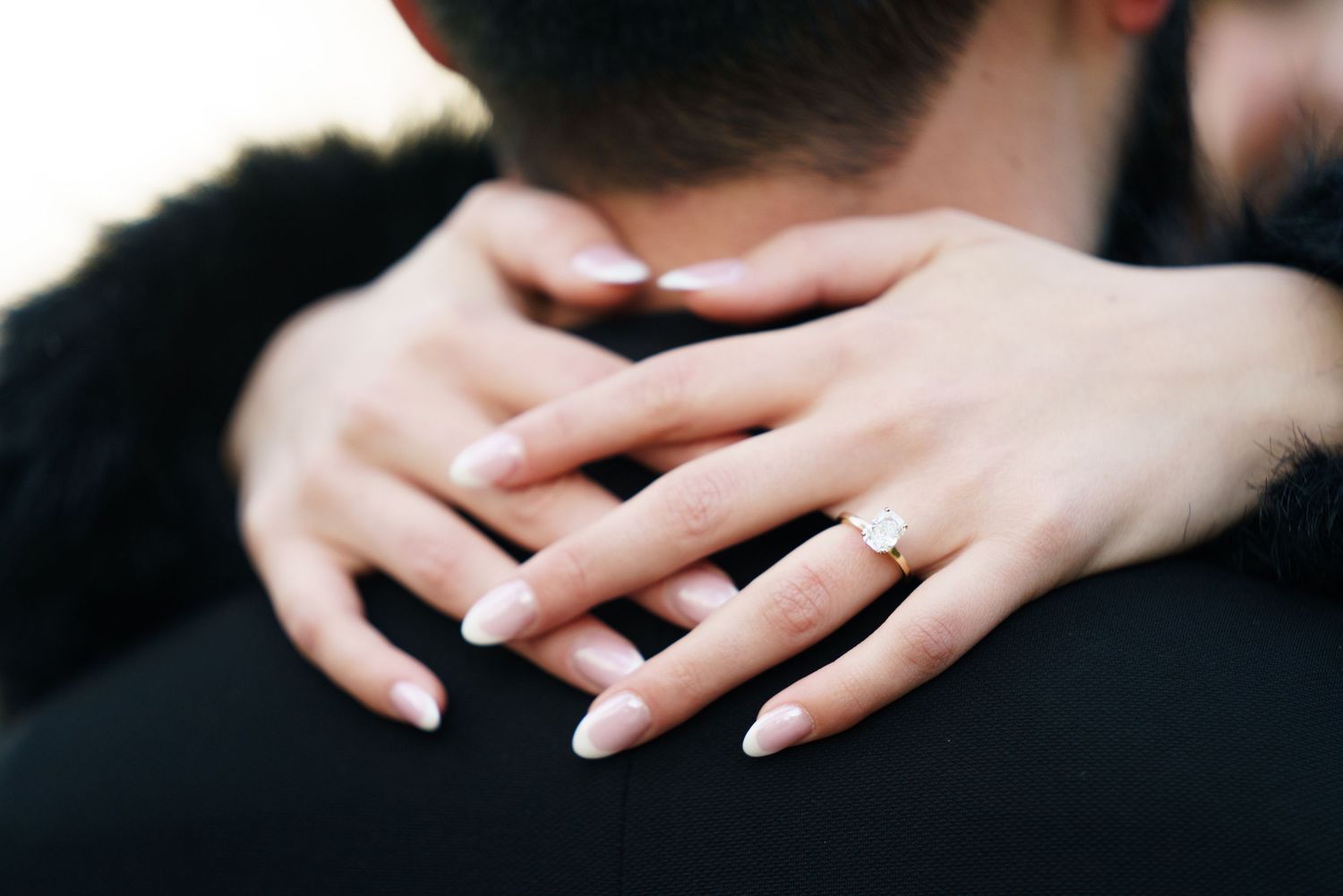 Close-up photo of hands wearing engagement rings against a dark background.