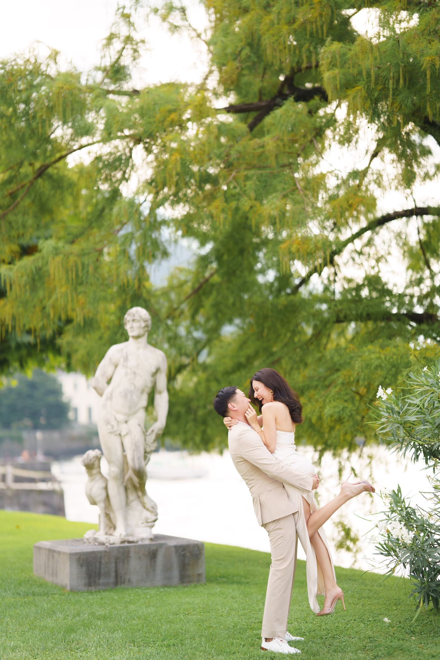 A romantic couple shares intimate moments in a garden setting with classical statues and lush greenery in the background.