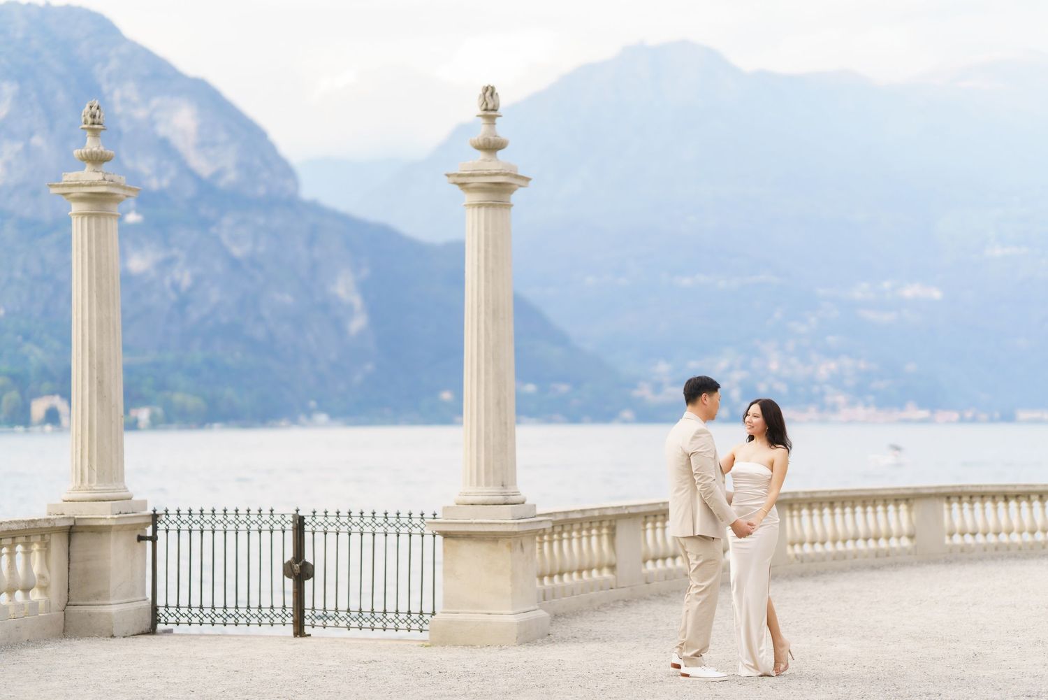 Couple in elegant attire embrace on a Mediterranean terrace overlooking coastal mountains and sea.
