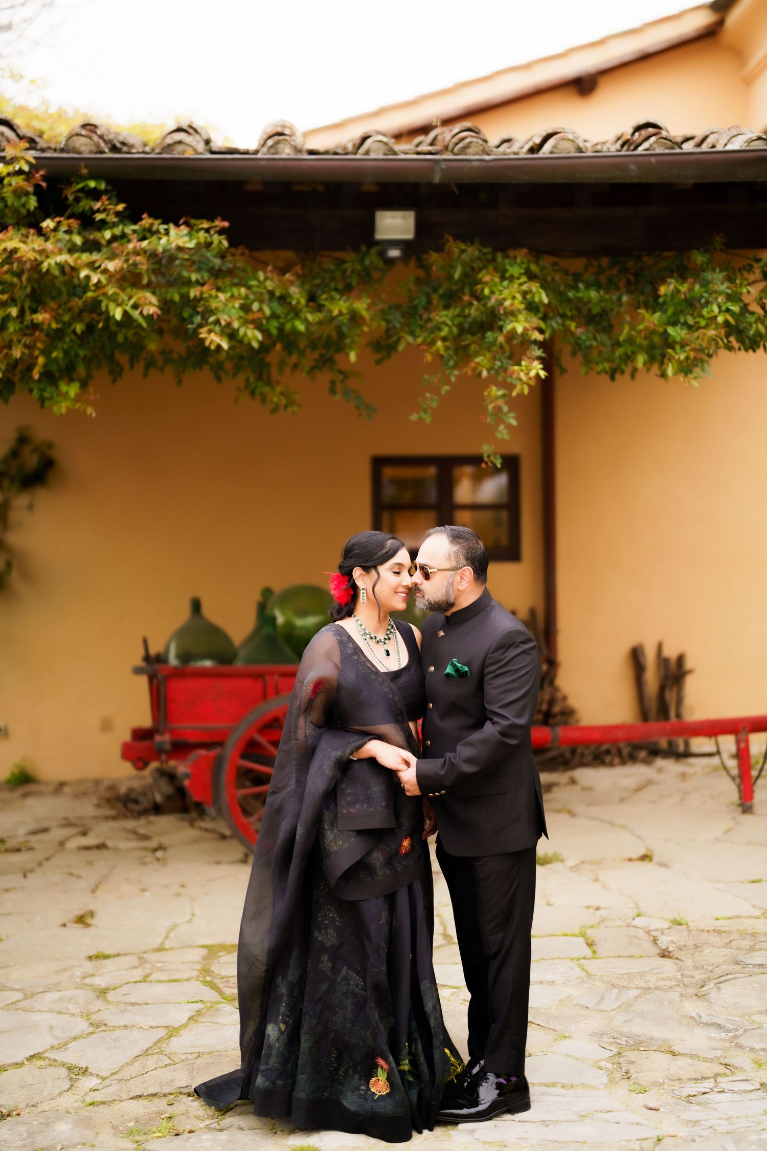 Romantic couple in formal attire sharing tender moment against rustic stone wall with red bench.