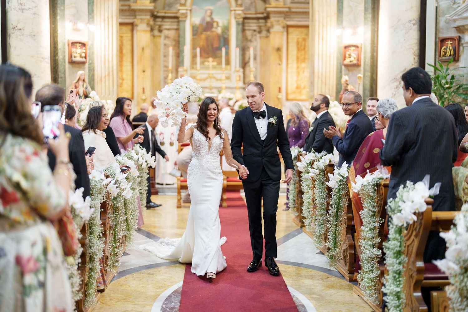 A wedding recessional in an ornate church with floral decorations lining the aisle.