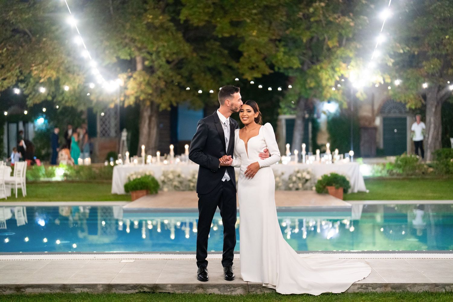 A nighttime wedding celebration by an illuminated pool with string lights overhead.