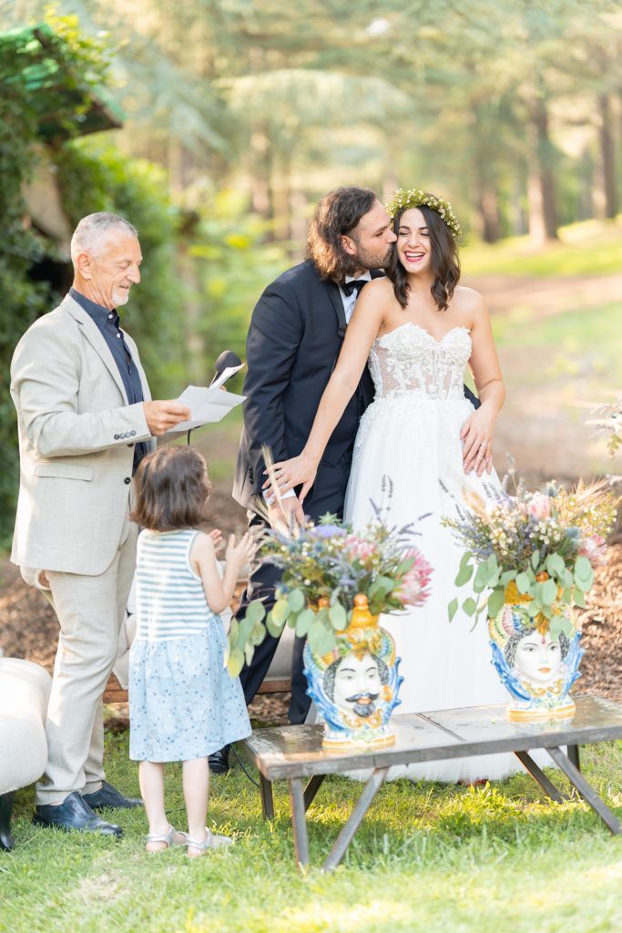 Wedding celebration with guests gathered around decorated table in sunny garden setting.