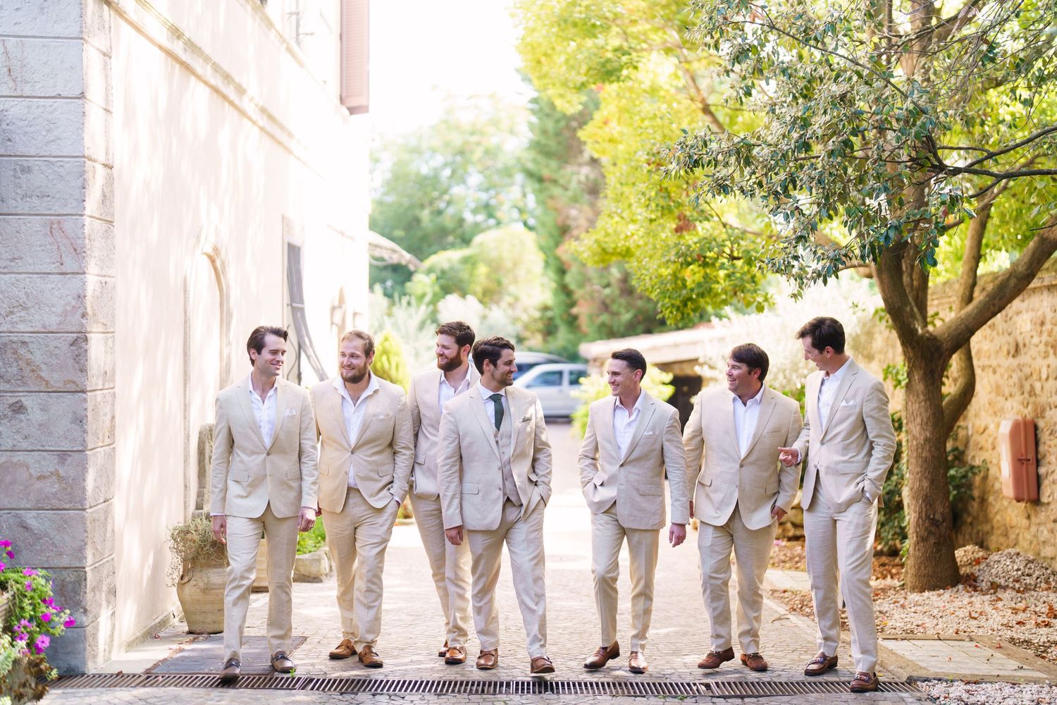 Groomsmen in light beige suits walking together along a stone path.
