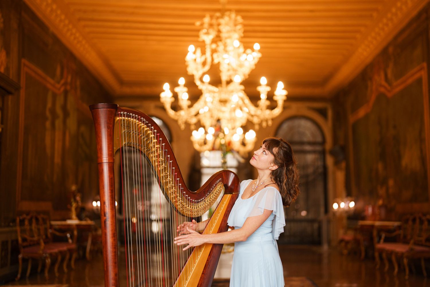 Elegant harpist in white dress performs in ornate historic hall with glowing crystal chandelier overhead.